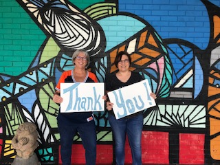 standing in front of a colorful mural two women hold signs that say thank and you. 