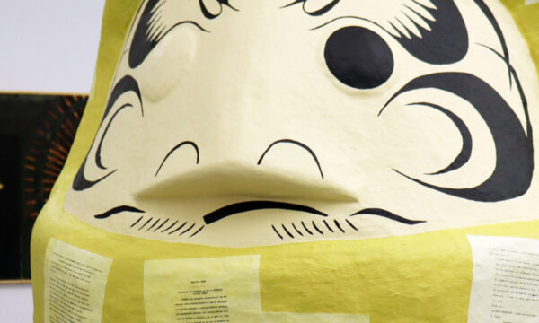 Large yellow sculpture with black markings that resemble a face with drawings on a gallery wall in the background.