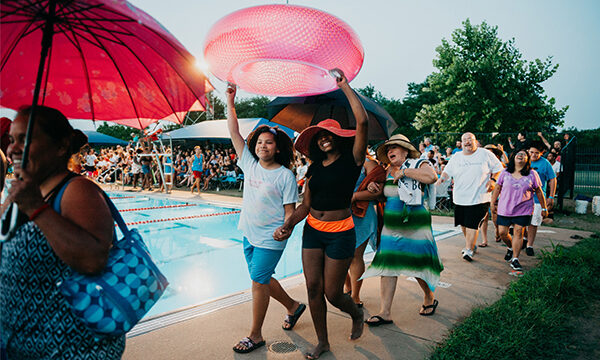People of all ages walk next to a pool in an art performance holding up floats.