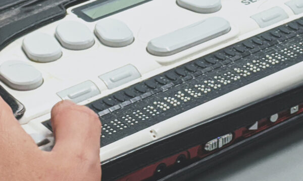 The hand of a person sitting at a desk uses a digital technology device with white keys and raised bumps to help the blind.