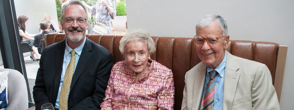 Man with suit on left smiles with two elderly individuals: one woman with red suit, a man with tan suit.