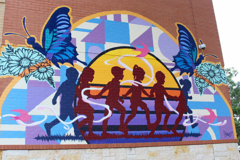 A mural on the side of a large brick building featuring silhouettes of children running with a colorful sunset and large purple and blue butterflies and abstracted designs.