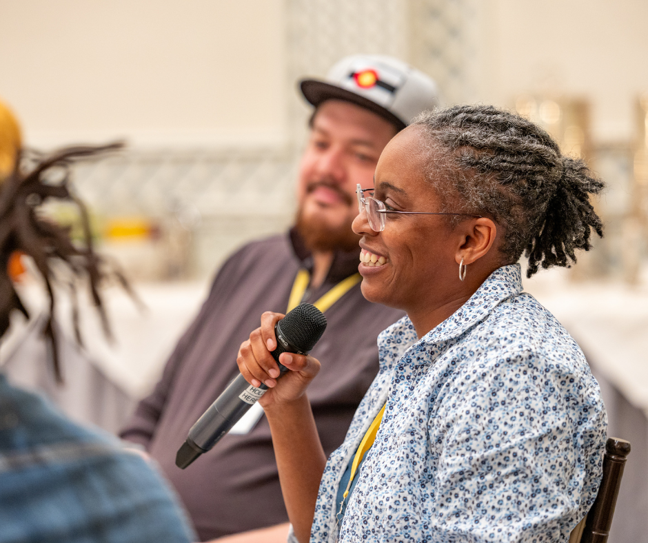 Black woman wearing glasses smiles and speaks into a microphone with people seated to either side of her, listening and smiling.