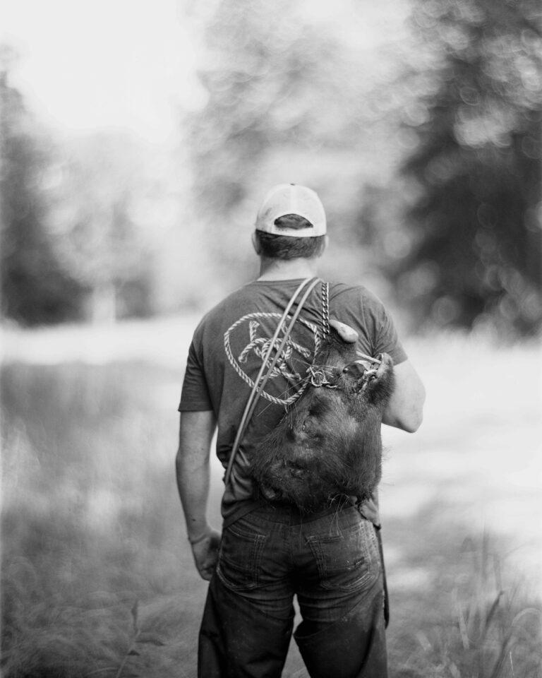 A man holds the head of a feral hog over his shoulder with his back to us in the woods.