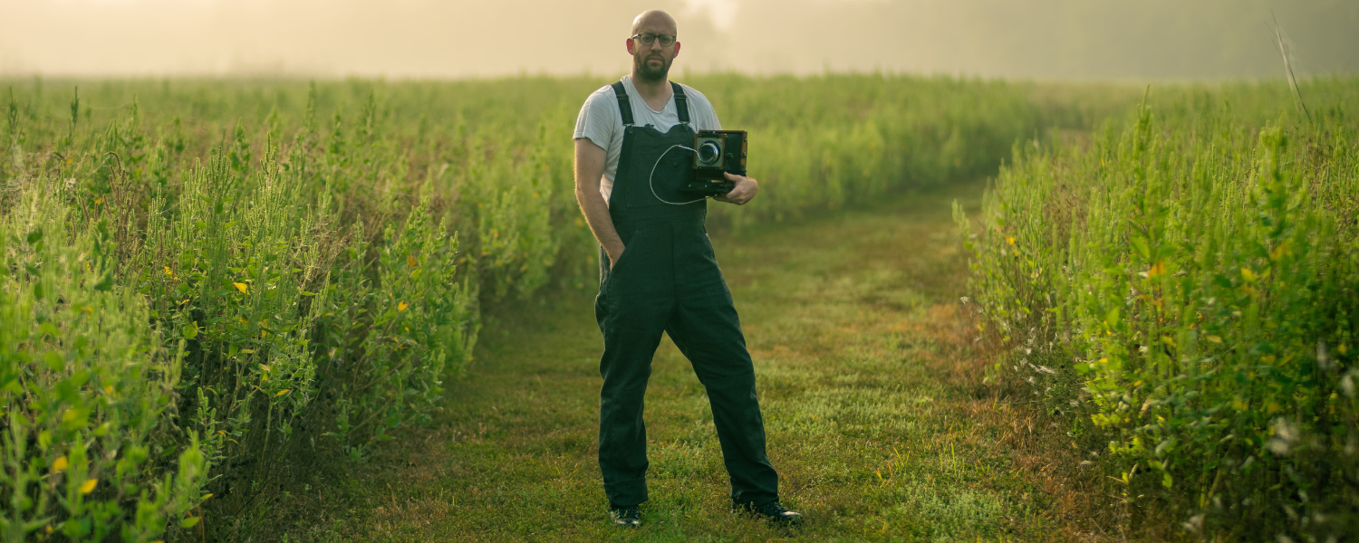 White man wearing overalls standing in a misty field holding a large format camera.