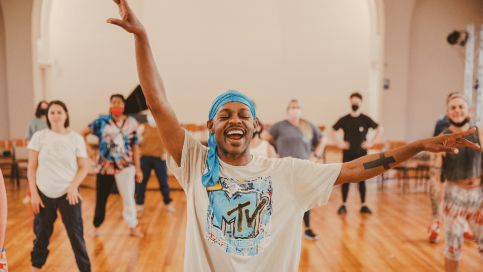 Black man smiles holding his arms up ready to lead a dance class with people behind him getting ready.