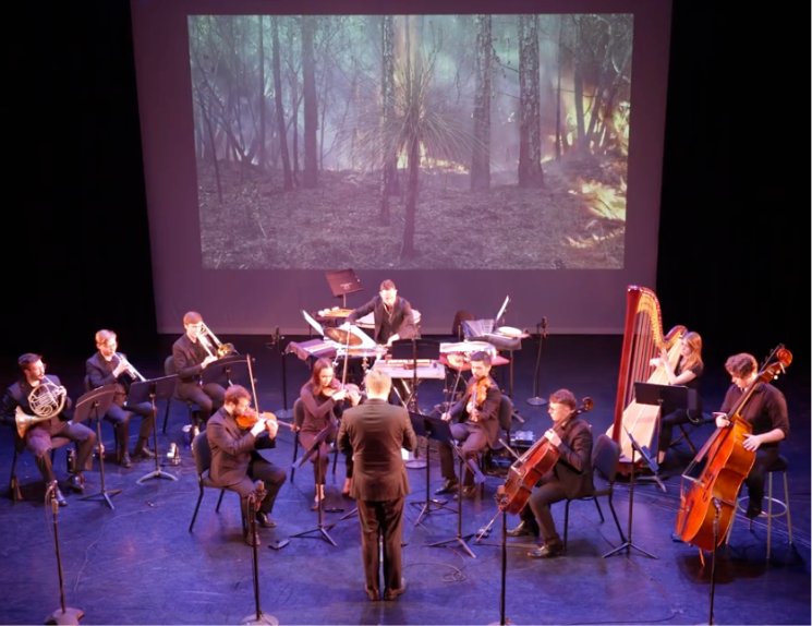 Ten musicians with a conductor perform onstage with a video projection behind them showing trees in a prescribed burn with flames and smoke seen in the video.