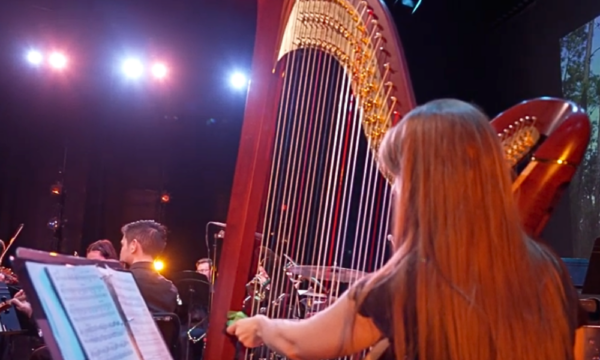 Woman plays a harp onstage with a musical ensemble and a video projection of a pine forest seen in the background.