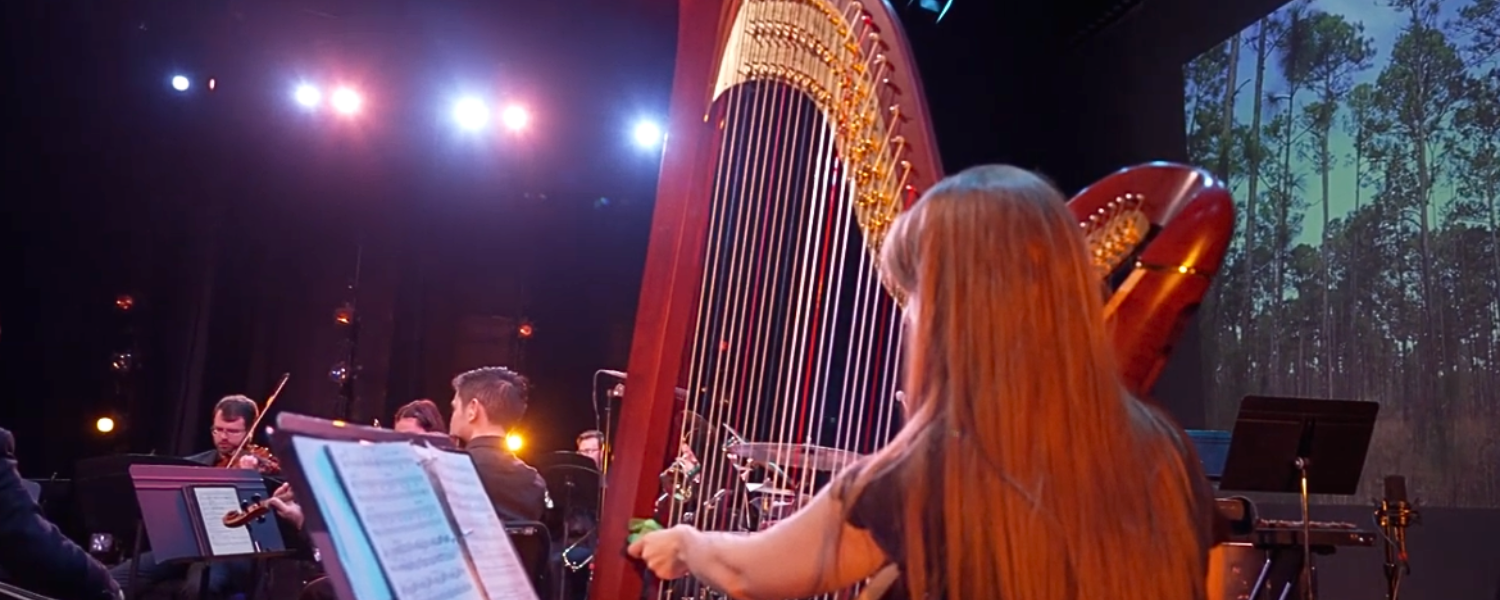 Woman plays a harp onstage with a musical ensemble and a video projection of a pine forest seen in the background.