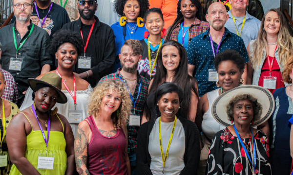 A large group of artists wearing brightly colored expressive clothing stand together on the stairs for a group portrait.