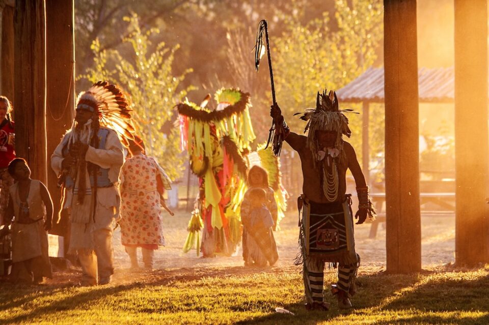 A Native American man in traditional costuming walks with other men, women, and children in a outdoor scene during sunset.