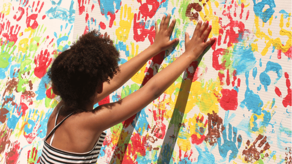 Young woman of color places her handprint on a wall of handprints in blue, red, yellow, and green paint colors.