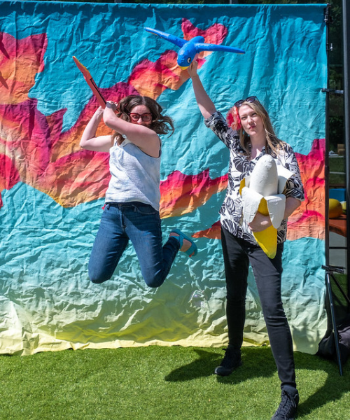 Woman jumping in the air wearing glasses and woman standing next to hear holding stuffed animals.