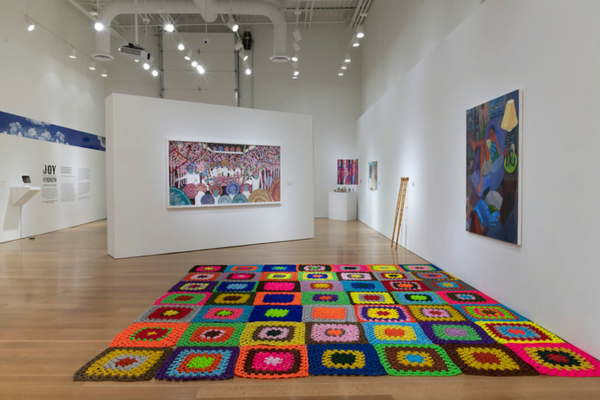 Large colorful blanket is spread out flat on a gallery floor with bright, joyful artworks in the background on large walls.