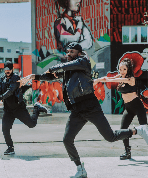 Men and women of color and diverse heritage dance hip hop in a public square outdoors with a mural behind them.