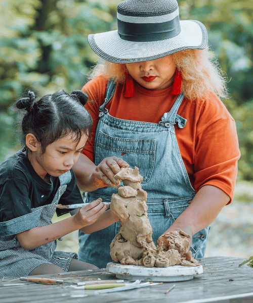 Woman of color wearing a brimmed hat assists a child making pottery in an outdoor setting.