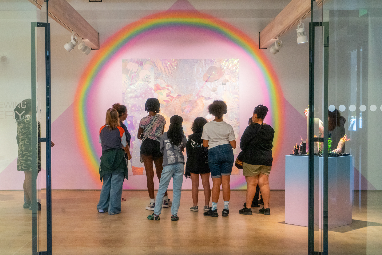 Nine youth and one instructor stand with their backs to the camera in front of a large, brightly-colored installation of foil and rainbow lights.