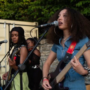 Three young woman of color play instruments and sing into a microphone on an outdoor stage.