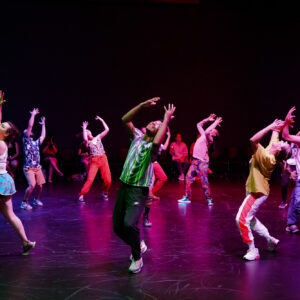 A group of 12 young adults dance in a circular formation raising hands and smiling in the middle of a dance movement onstage. 