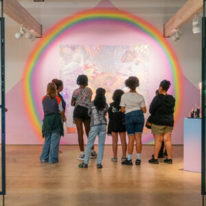 Nine youth and one instructor stand with their backs to the camera in front of a large, brightly-colored installation of foil and rainbow lights.