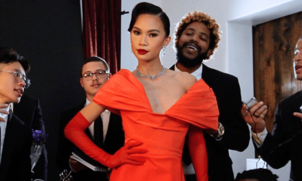 Woman stands in a glamorous formal red dress with men in suits looking at her attentively.