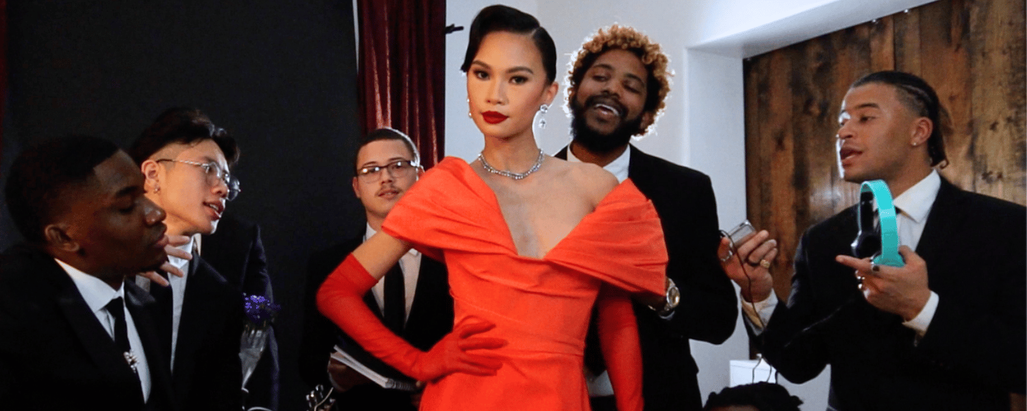 Woman stands in a glamorous formal red dress with men in suits looking at her attentively.