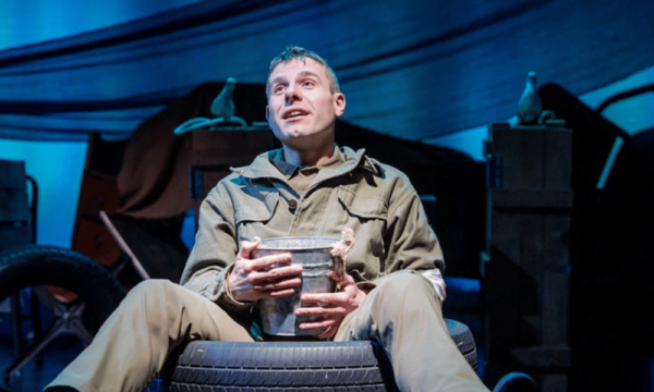 Man onstage in a military setting wears a green uniform and sits on a tire looking up to the sky.