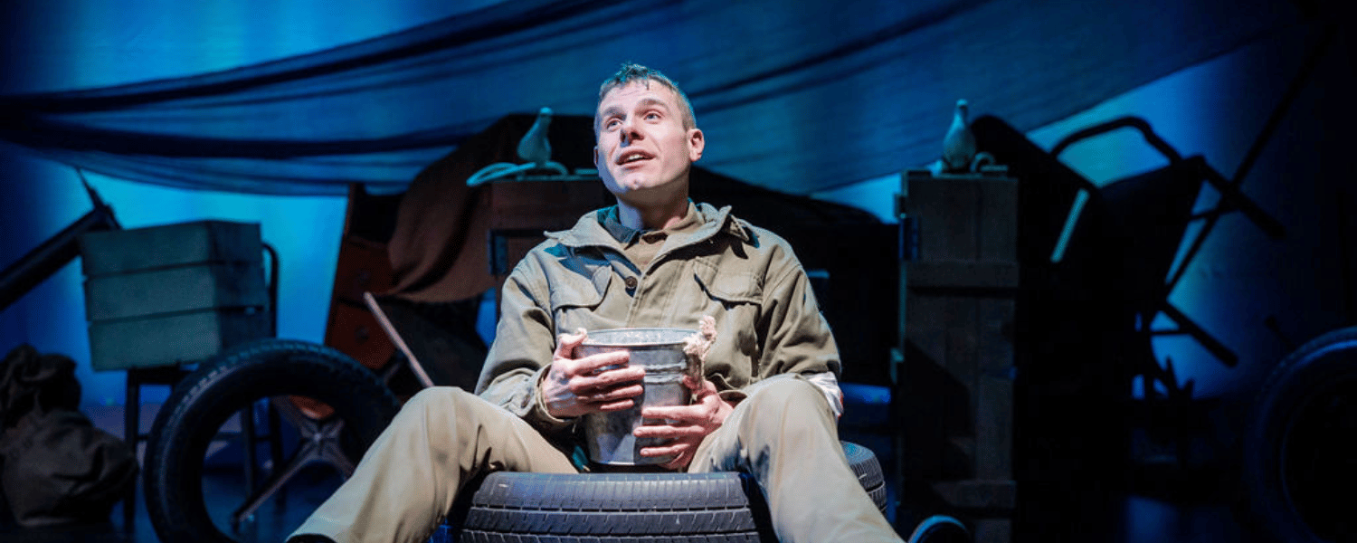 Man onstage in a military setting wears a green uniform and sits on a tire looking up to the sky.