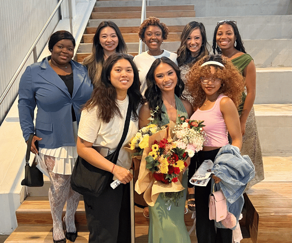 Group of artists pose together around one artist with flowers.