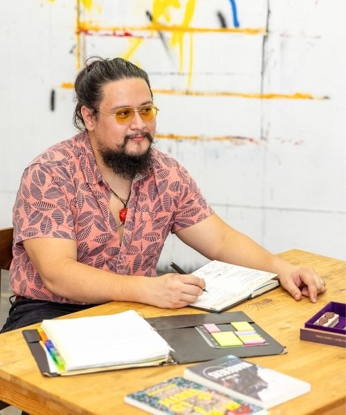 Man with a beard sits at a table with a notebook in an art studio.