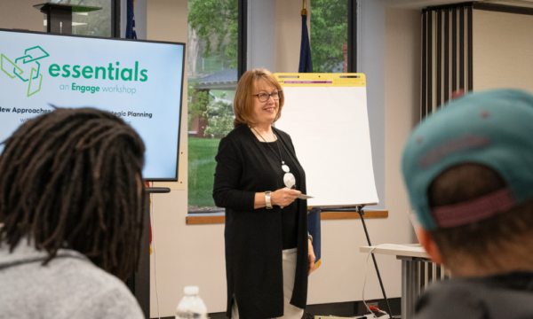 Smiling woman stands in facilitates discussion for a group in a classroom setting.