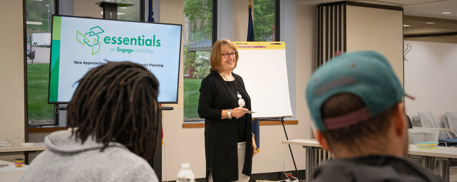 Smiling woman stands in facilitates discussion for a group in a classroom setting.