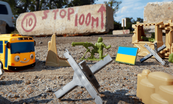 A plastic yellow school bus, plastic toy jacks, a plastic Ukrainian flag and two plastic green 'army men' solider toys are scattered on the ground. The two soldier toys aim their weapons at the bus as it approaches. In the background sits a concrete brick with the words 