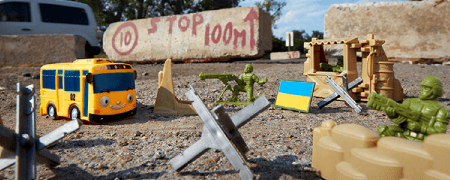 A plastic yellow school bus, plastic toy jacks, a plastic Ukrainian flag and two plastic green 'army men' solider toys are scattered on the ground. The two soldier toys aim their weapons at the bus as it approaches. In the background sits a concrete brick with the words 