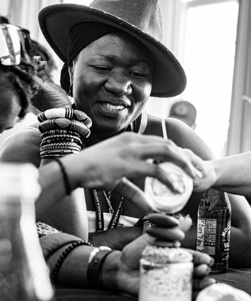 A Black female-presenting individual sits at a table with many people pouring sand and stones into a cup.