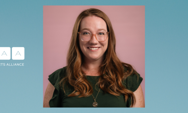 A woman with long reddish brown hair glasses and a dark green shirt smiles for the camera in a headshot portrait photo.