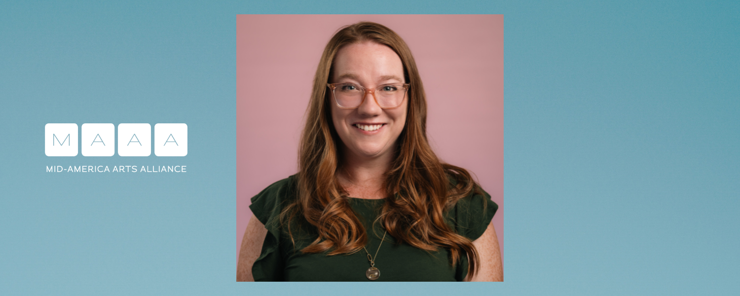 A woman with long reddish brown hair glasses and a dark green shirt smiles for the camera in a headshot portrait photo.