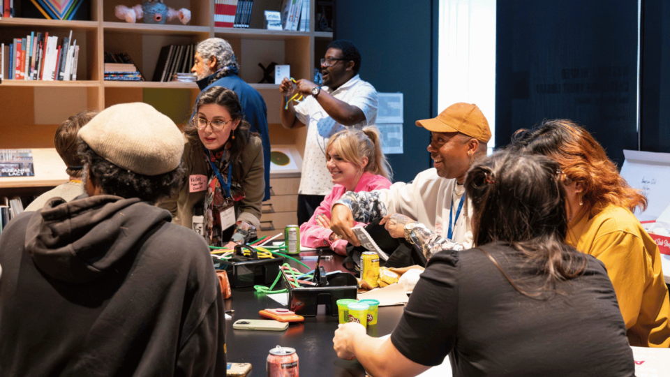 Seven artists sit around a table talking and smiling. 