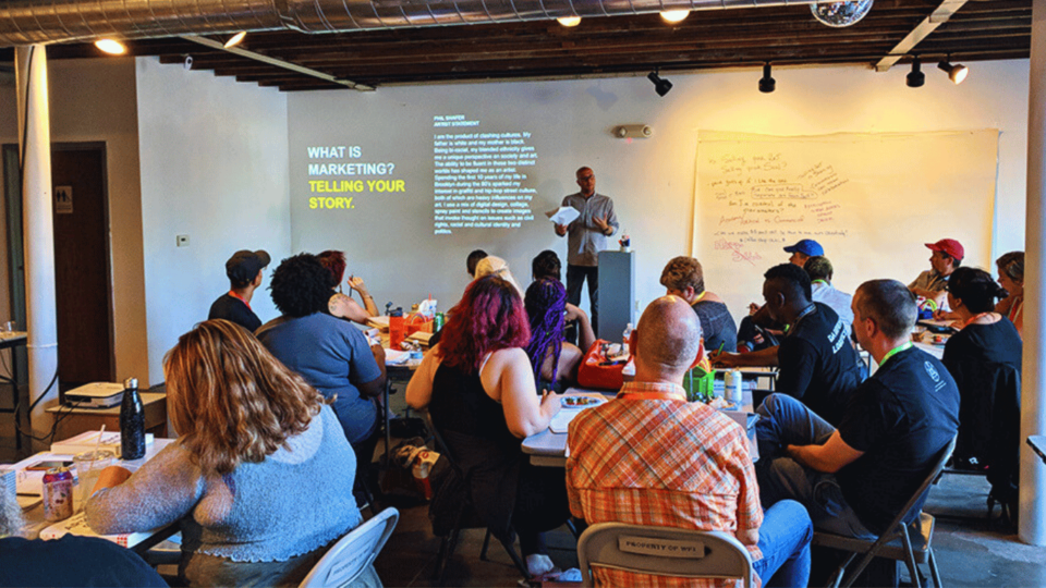 A man speaks to a classroom of creatives sitting at tables with notebooks and fun activities.