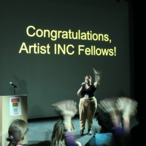 A woman holds a microphone in a dark theatrical setting and waves with hands clapping in the foreground. The projection behind her says Congratulations Artist INC Fellows.