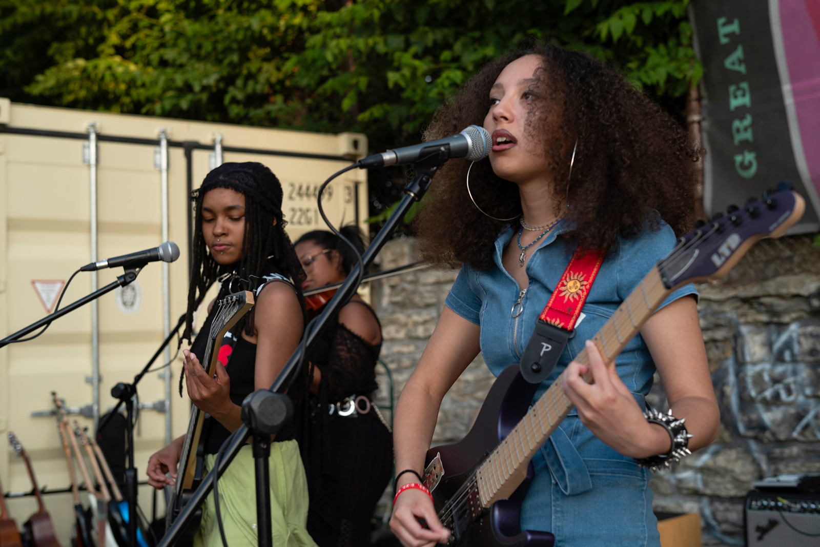 Three young women of color play guitars and a violin and sing into microphones at an outdoor concert.