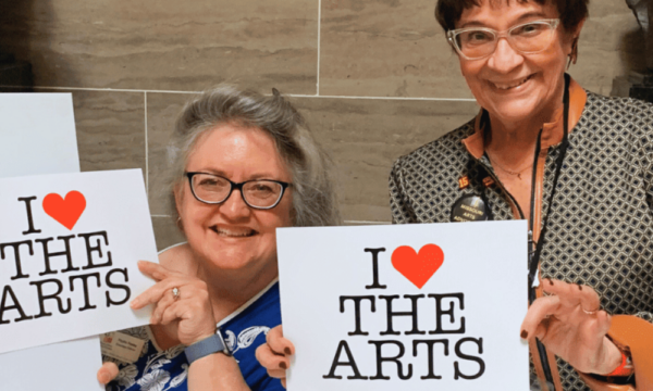 Two smiling women hold up signs that say I Heart the Arts.