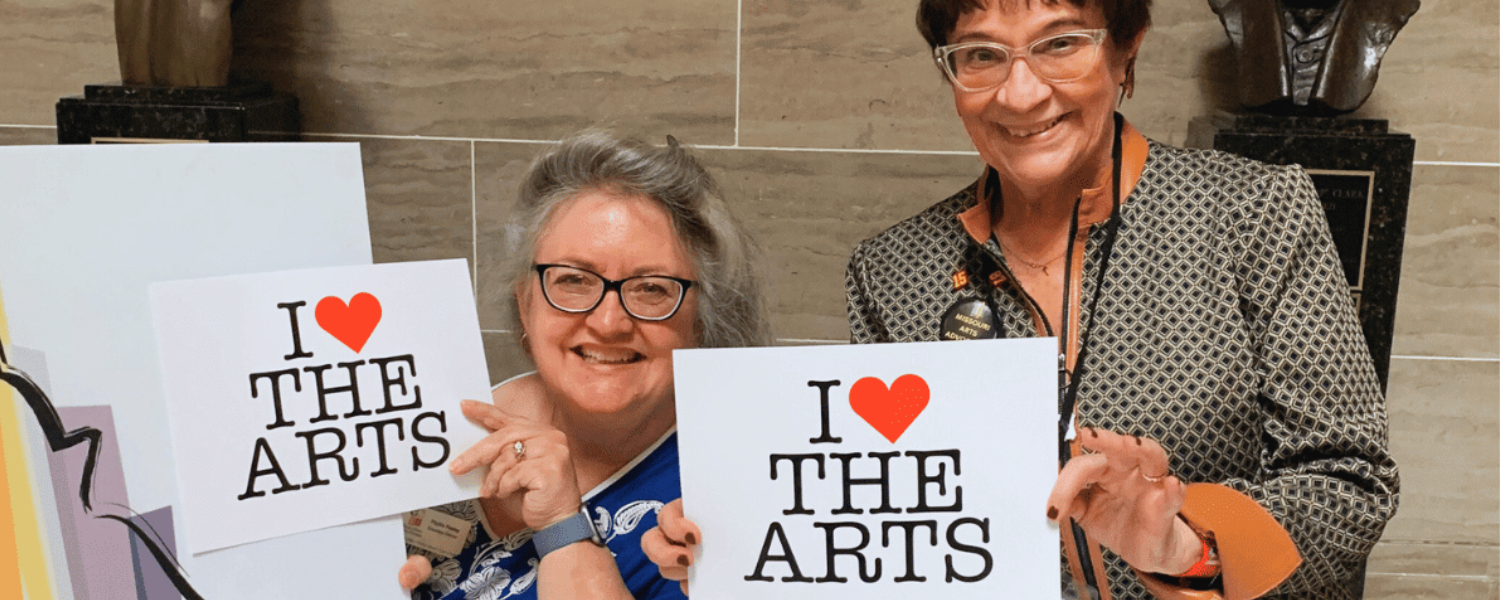 Two smiling women hold up signs that say I Heart the Arts.