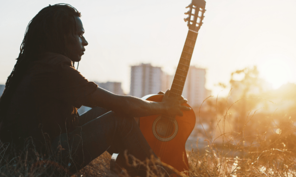 A Black man sits on a grass hill with his acoustic guitar looking over a cityscape and sunset in the background.