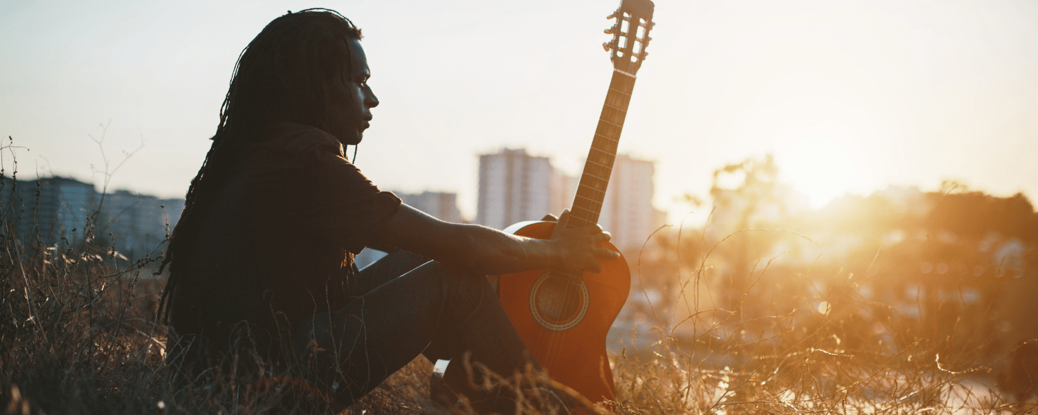 A Black man sits on a grass hill with his acoustic guitar looking over a cityscape and sunset in the background.