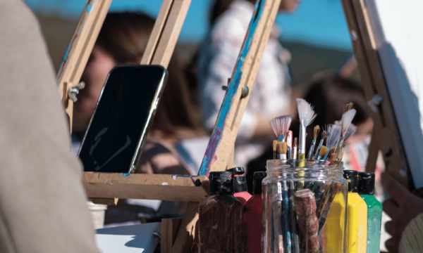 People sit around a table of easels, canvases, and a large glass jar of paintbrushes. One easel holds a smartphone.