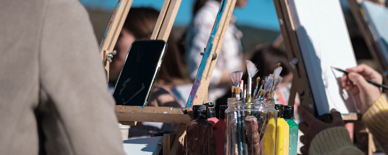 People sit around a table of easels, canvases, and a large glass jar of paintbrushes. One easel holds a smartphone.