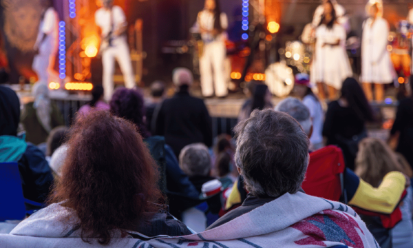 A large community gathering outdoors with lawn chairs and blankets for an outdoor concert with many musicians, lights, and instruments onstage.