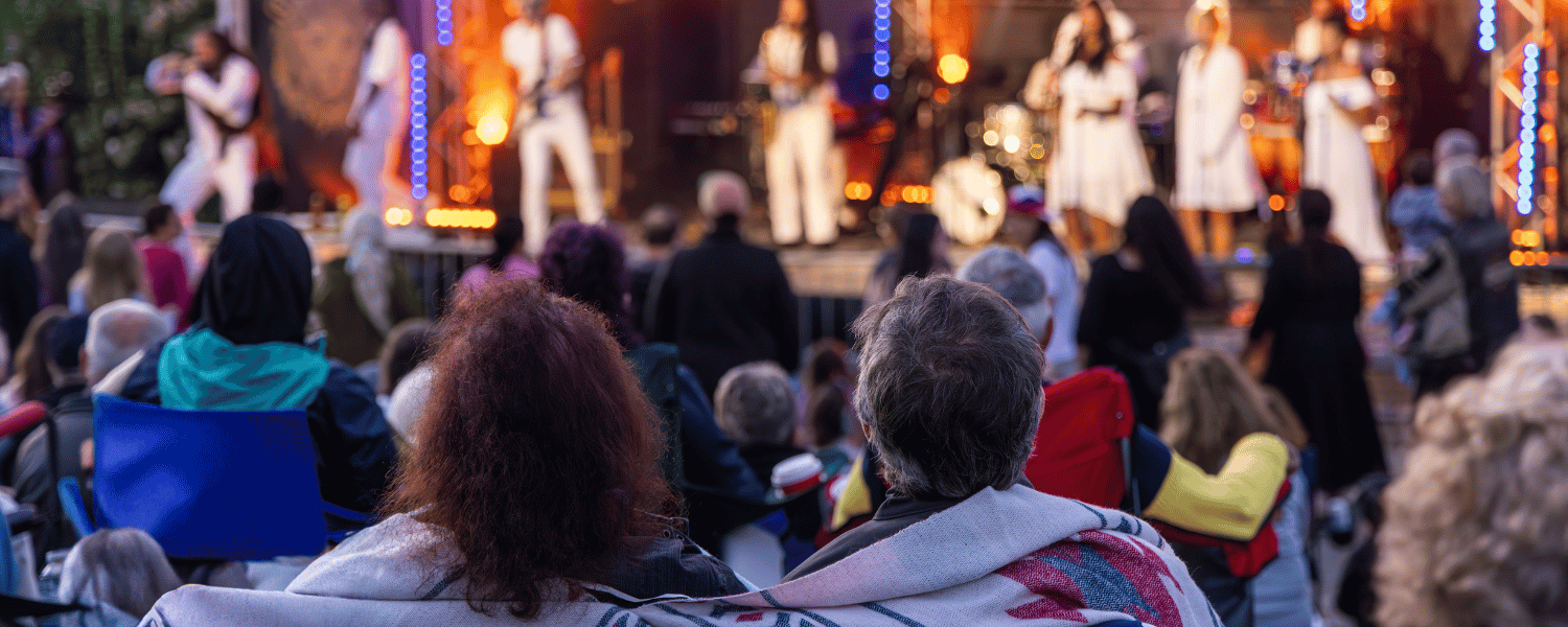 A large community gathering outdoors with lawn chairs and blankets for an outdoor concert with many musicians, lights, and instruments onstage.