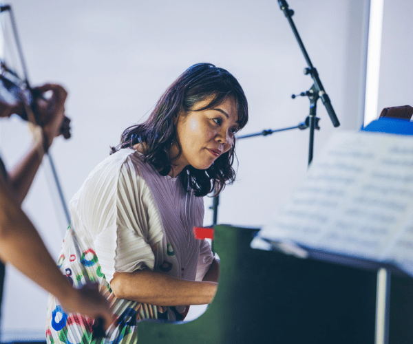 An asian woman plays the piano with a person playing a violin in the foreground.